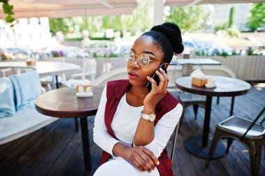 Afro-Amerikan Kız giyim gözlük ile hareket eden telefon açık caffe oturan.