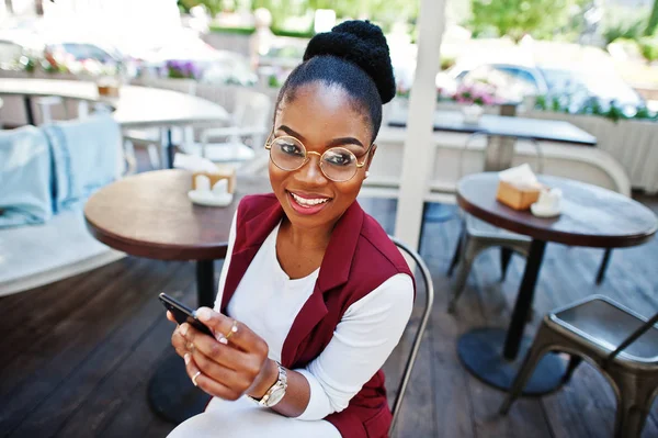 Afro-Amerikan Kız giyim gözlük ile hareket eden telefon açık caffe oturan.