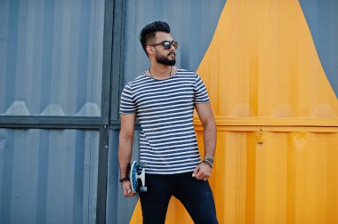 Yakışıklı uzun boylu Arap sakal man modeli soyulmuş gömlek, açık poz verdi. Sarı boyalı duvar karşı kaykay ile güneş gözlüğü moda Arap adama.