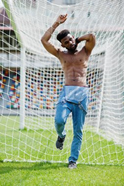 Yakışıklı seksi Afro-Amerikan kaslı çıplak gövde erkek kot Tulum, yeşil çim agasinst futbol kapılarında Stadyumu alanının poz verdi. Şık siyah adam portresi.