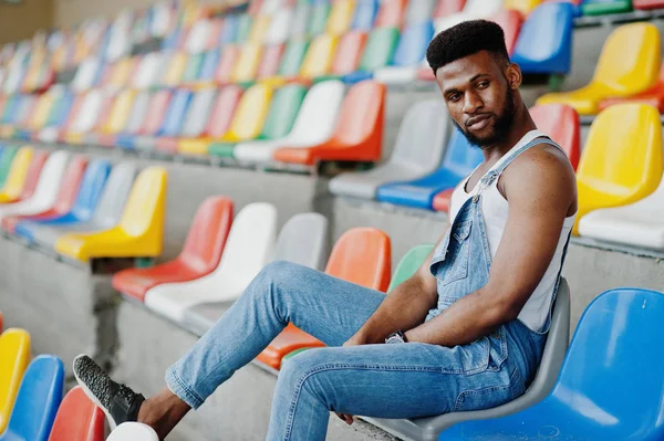 Handsome african american man at jeans overalls posed on colored chairs at stadium. Fashionable black man portrait.