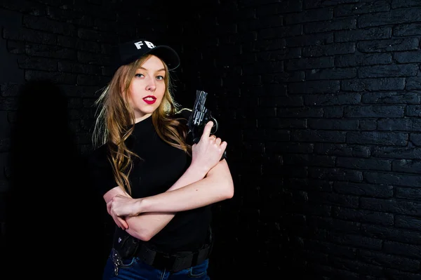 FBI female agent in cap and with gun at studio against dark brick wall.