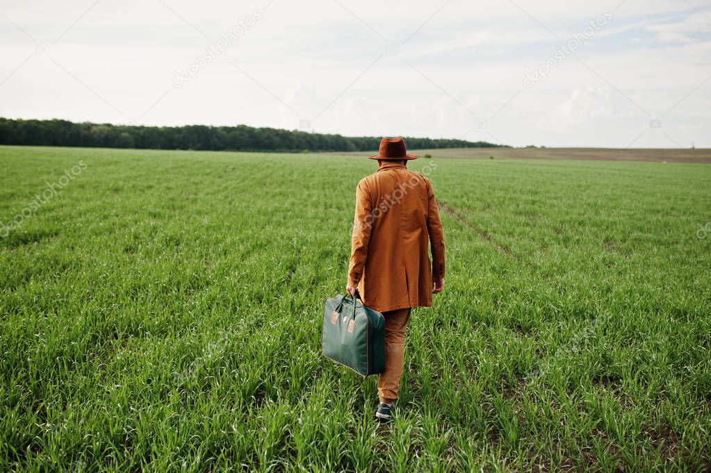 Stylish man in glasses, brown jacket and hat with bag posed on green field.