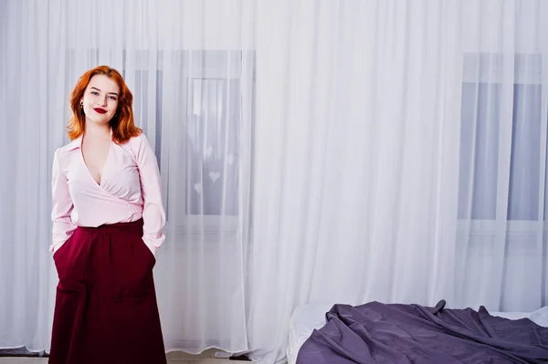 Portrait of a beautiful redheaded girl in blouse and skirt standing by the bed.