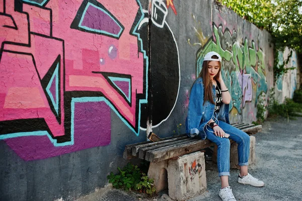 时尚休闲时髦的女孩在帽子和牛仔裤穿着听音乐从手机耳机对大涂鸦墙与炸弹 — 图库照片