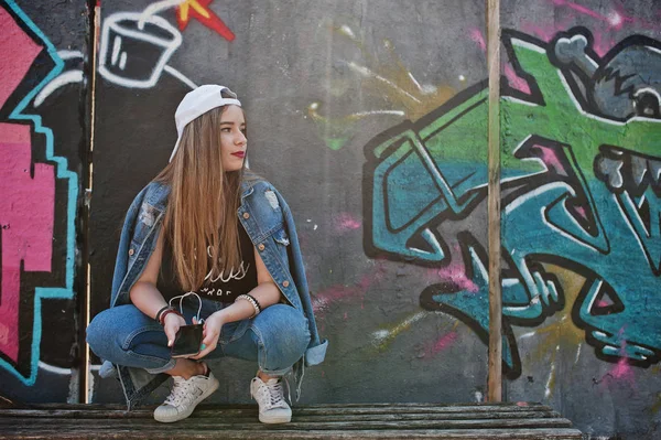 时尚休闲时髦的女孩在帽子和牛仔裤穿着听音乐从手机耳机对大涂鸦墙与炸弹 — 图库照片