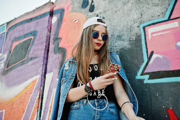 时尚的休闲时髦女孩帽 太阳镜和牛仔裤穿 听音乐从手机耳机对大涂鸦墙 — 图库照片