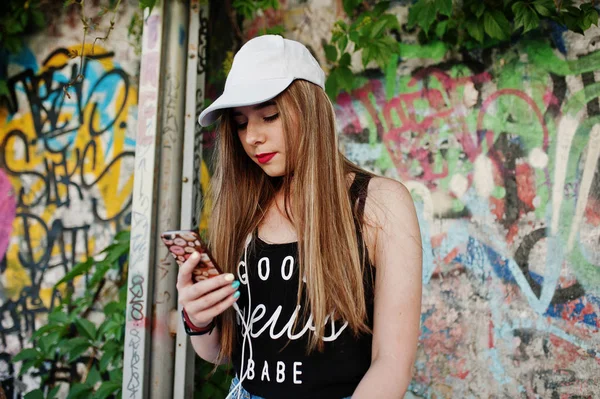 大きな落書きの壁に対して携帯電話のヘッドフォンから音楽を聴くキャップ ジーンズ着用でスタイリッシュなカジュアルな内気な少女 — ストック写真