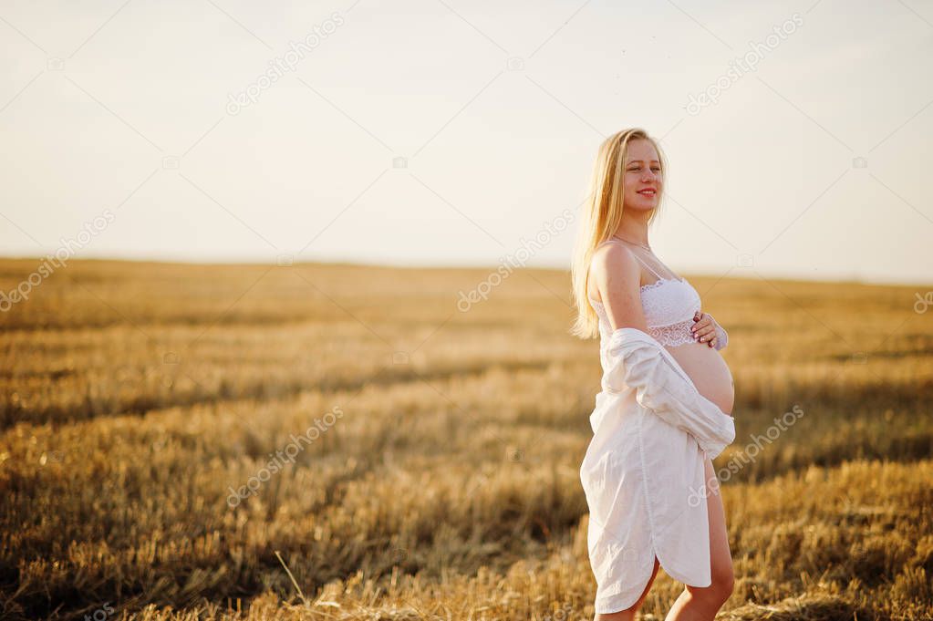 Blonde pregnant woman in wreath field at white underwear clothes on sunset. Happy moments of pregnancy.