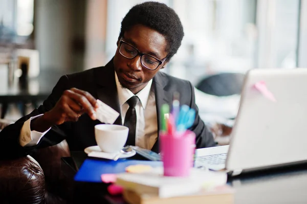 Business african american man wear on black suit and glasses sitting at office with laptop and working, pours sugar into coffee.