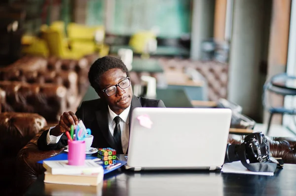 Business african american man wear on black suit and glasses sitting at office with laptop and working, pours sugar into coffee.