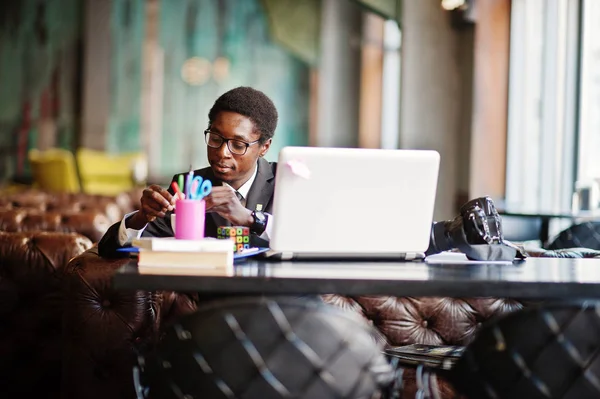 Business african american man wear on black suit and glasses sitting at office with laptop and working, pours sugar into coffee.