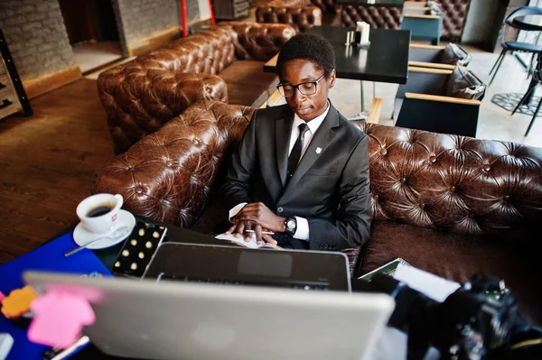 Business african american man wear on black suit and glasses sitting at office with laptop and working, reading magazine.