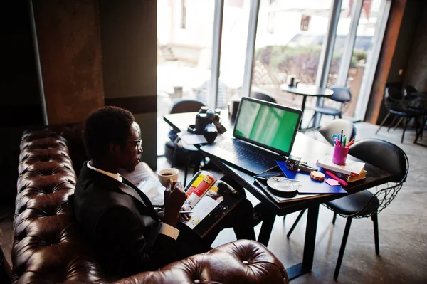 Business african american man wear on black suit and glasses sitting at office with green screen laptop and working, reading magazine and drinking coffee.