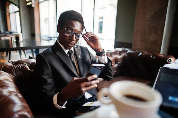 Business african american man wear on black suit and glasses sitting at office with laptop and working, looking at mobile phone.