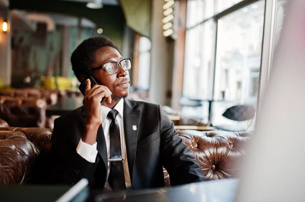 Business african american man wear on black suit and glasses sitting at office and speaking on phone behind laptop.