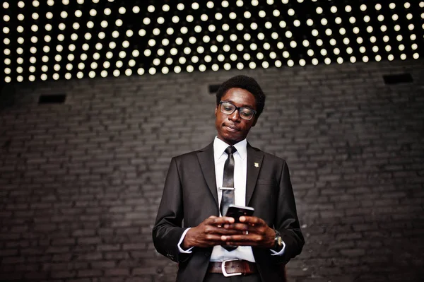 Business african american man wear on black suit and glasses at office looking on mobile phone against futuristic lights.