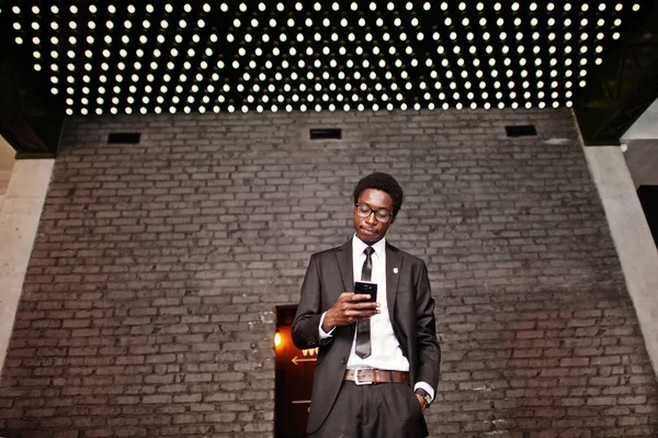 Business african american man wear on black suit and glasses at office looking on mobile phone against futuristic lights.