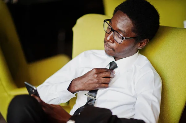 Business african american man wear on white shirt, tie and glasses at office, sitting on chair and holding mobile phone.