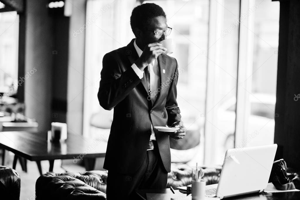 Business african american man wear on black suit and glasses at office with laptop, drinking morning coffee before work day.