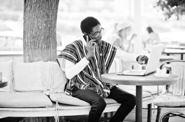 Geleneksel giysiler ve açık caffe laptop arkasında oturan ve cep telefonu konuşma gözlük Afrika adam.