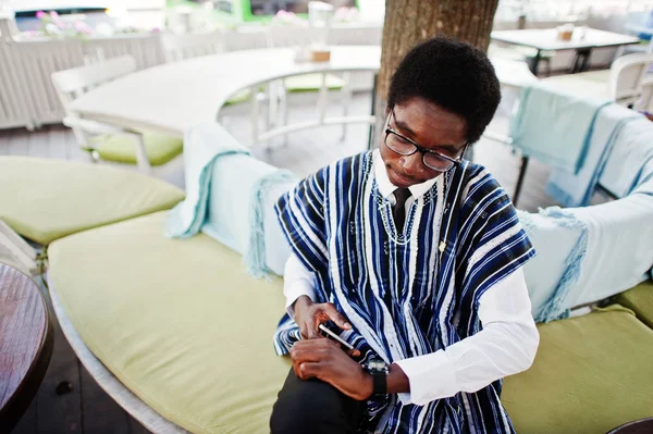 African man in traditional clothes and glasses sitting at outdoor caffe with cell phone.