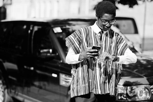 African businessman in traditional clothes and glasses with mobile phone against black car suv looking on his watches. Rich africans people.