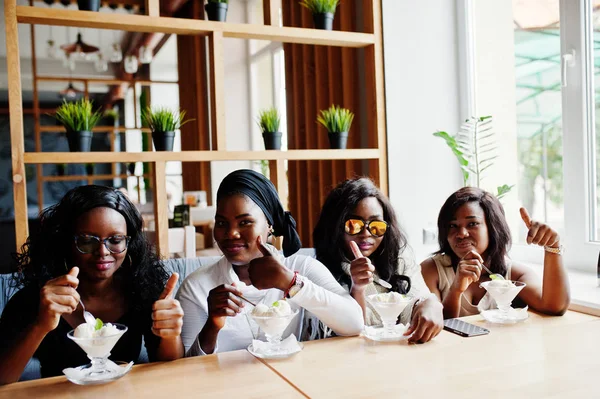 Four african american girls sitting on table at cafe and eating ice cream dessert and showing thumb up.