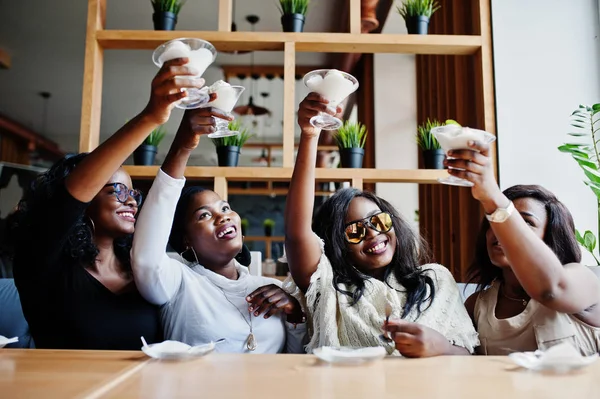 Four african american girls sitting on table at cafe and cheers with ice cream dessert.