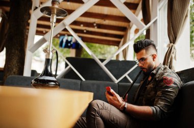 Arap adam gözlük ve askeri ceket sigara nargile sokak barında şık sakal. Dinlenme ve telefonla arıyor Arap modeli.