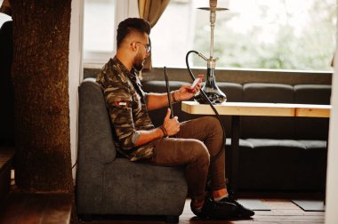 Arap adam gözlük ve askeri ceket sigara nargile sokak barında şık sakal. Dinlenme ve alma selfie telefonla Arap modelinde.