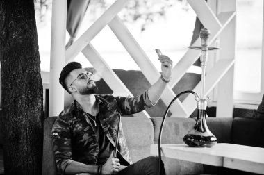Arap adam gözlük ve askeri ceket sigara nargile sokak barında şık sakal. Dinlenme ve alma selfie telefonla Arap modelinde.