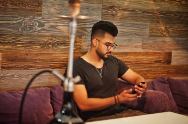 Arap adam gözlük ve siyah tişört sigara nargile bar kapalı şık sakal. Dinlenme ve seyir telefon Arap modeli.