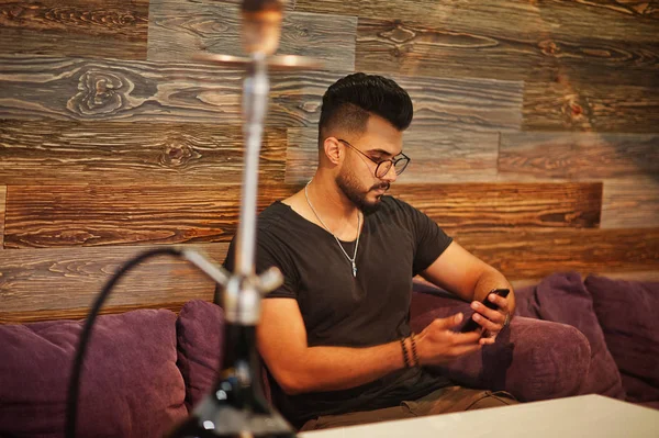 Arap adam gözlük ve siyah tişört sigara nargile bar kapalı şık sakal. Dinlenme ve seyir telefon Arap modeli.