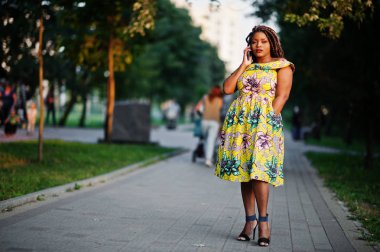 Sevimli küçük yükseklik dreadlocks, Afrikalı-Amerikalı kızla giymek batımında, poz renkli sarı elbise yürüyüş ve cep telefonunuzun konuşma.