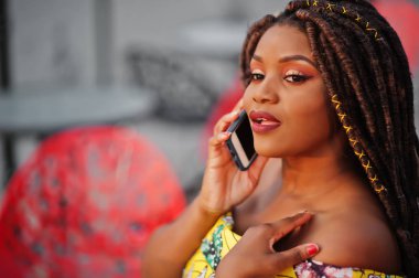 Sevimli küçük yükseklik dreadlocks, Afrikalı-Amerikalı kızla açık kafede kırmızı sandalye üzerinde oturan ve konuşan renkli sarı elbise giymek.