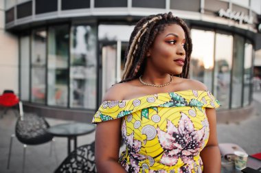 Sevimli küçük yükseklik Afro-Amerikan Kız dreadlocks, bina büyük pencereler karşı yöneltilen renkli sarı elbise giymek.