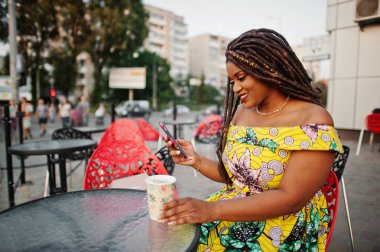 Sevimli küçük yükseklik dreadlocks, Afrikalı-Amerikalı kızla giymek açık kafede kırmızı sandalye üzerinde oturan ve içme kahve renkli sarı elbise,.