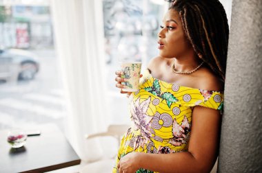 Sevimli küçük yükseklik dreadlocks, Afrikalı-Amerikalı kızla kafede kahve ile poz renkli sarı elbise giymek.
