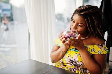 Sevimli küçük yükseklik Afro-Amerikan Kız dreadlocks, çiçek, cam ile kafede oturan renkli sarı elbise giymek.