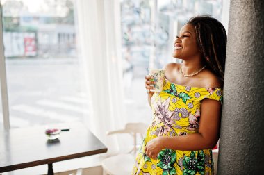 Sevimli küçük yükseklik dreadlocks, Afrikalı-Amerikalı kızla kafede kahve ile poz renkli sarı elbise giymek.