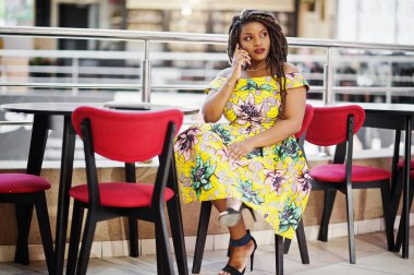 Sevimli küçük yükseklik Afro-Amerikan Kız dreadlocks, Alışveriş Merkezi kafede oturan ve konuşan renkli sarı elbise giymek.