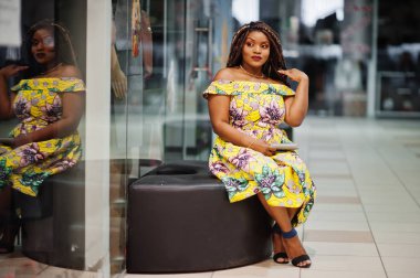 Sevimli küçük yükseklik Afro-Amerikan Kız dreadlocks, alışveriş merkezinde bankta oturmuş renkli sarı elbise giymek.