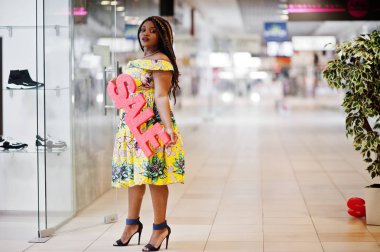 Sevimli küçük yükseklik dreadlocks, Afrikalı-Amerikalı kızla giymek renkli sarı elbise, Satılık işareti mektup alışveriş merkezi üzerinde tutan.