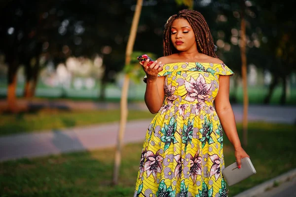 Sevimli küçük yükseklik dreadlocks, Afrikalı-Amerikalı kızla giymek batımında, poz renkli sarı elbise seyir telefon.