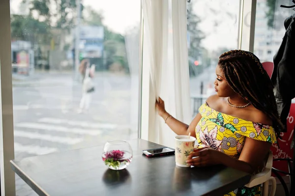 Sevimli küçük yükseklik dreadlocks, Afrikalı-Amerikalı kızla kafede kahve fincan ile oturan ve penceresine bakarak renkli sarı elbise giymek.