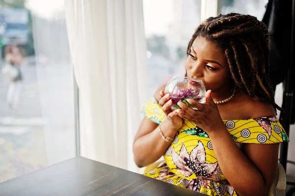 Sevimli küçük yükseklik Afro-Amerikan Kız dreadlocks, çiçek, cam ile kafede oturan renkli sarı elbise giymek.