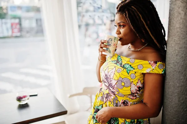 Sevimli küçük yükseklik dreadlocks, Afrikalı-Amerikalı kızla kafede kahve ile poz renkli sarı elbise giymek.