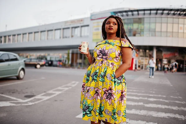 Cute small height african american girl with dreadlocks, wear at coloured yellow dress, walking at crosswalk against trade center with cup of coffee at hand.