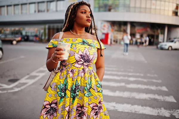 Cute small height african american girl with dreadlocks, wear at coloured yellow dress, walking at crosswalk against trade center with cup of coffee at hand.